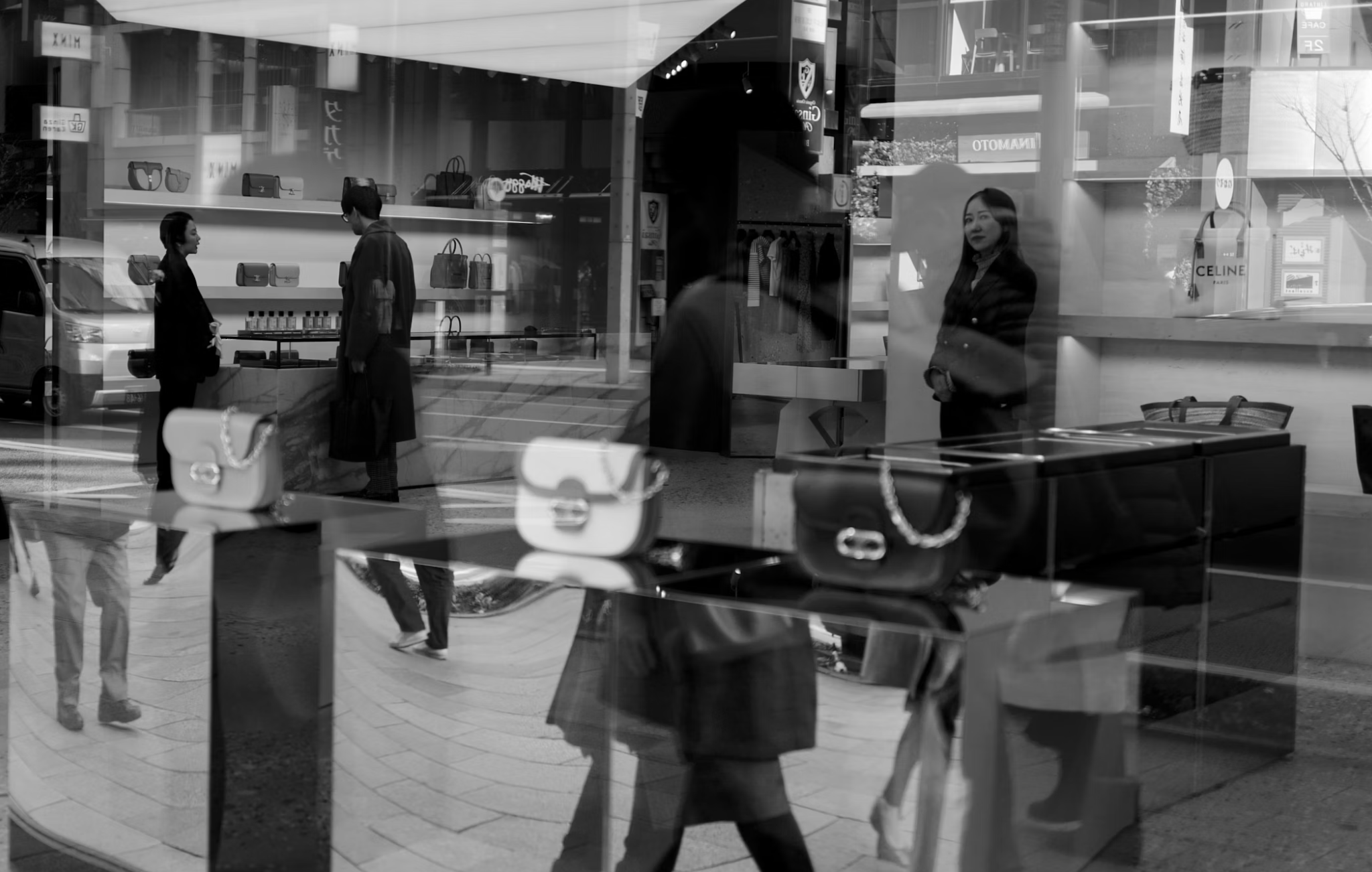 Black-and-white photo of a luxury store window with handbags and layered street reflections.